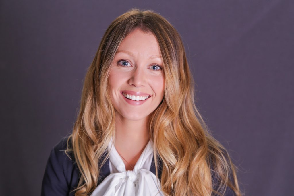 Corporate looking woman with blonde hair smiling into the camera wearing white blouse with a bow