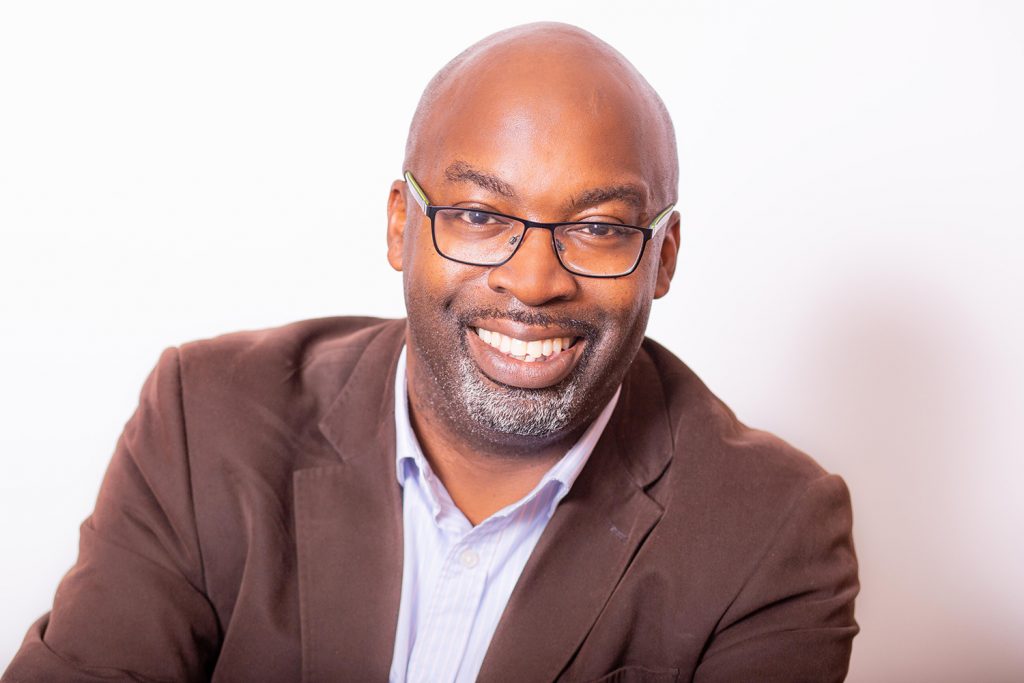 Friendly black man with glasses and brown jacket smiling.