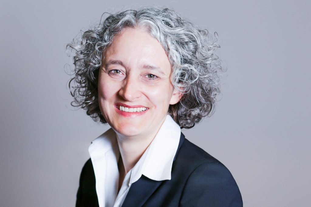 Corporate lady with grey curly hair smiling into the camera in front of grey backdrop