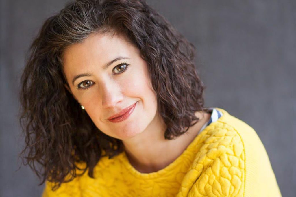 Woman with black curly hair staring into camera wearing bright yellow top
