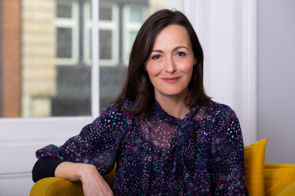 Gentle looking woman with lovely smile sitting on mustard coloured chair in front of window