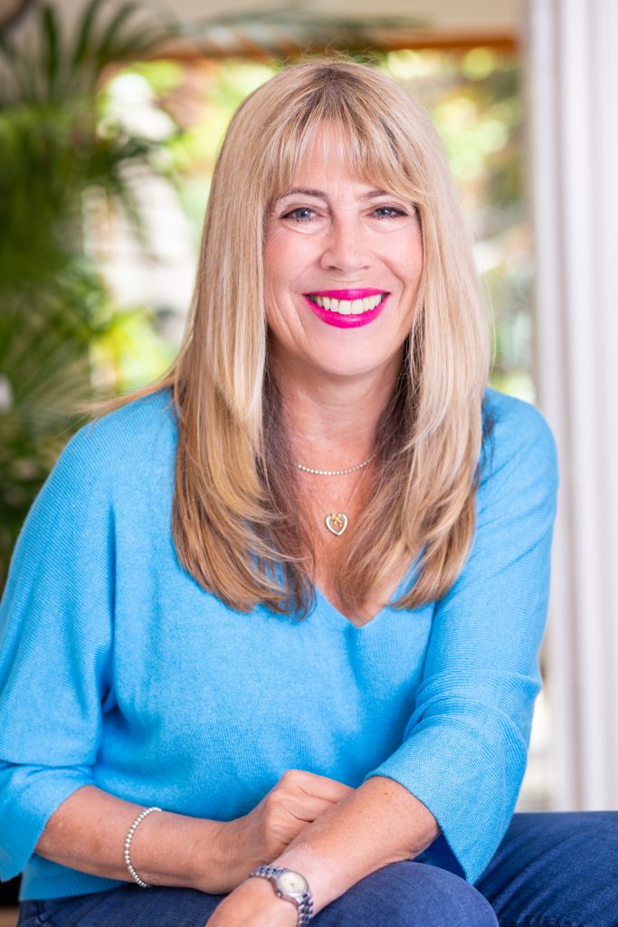 Blonde woman smiling at camera with garden in the background. Wears bright turquoise shirt and jeans