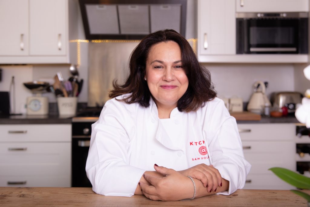 Friendly cheerful woman chef sitting at kitchen table in front of oven and kitchen utensils. Wears chef's coat