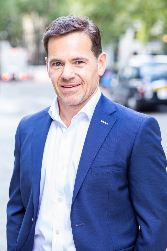 Corporate looking man in blue suit outside in the street with cars in the background and smiling into the camera